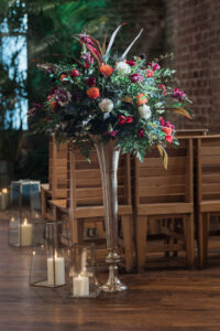 Silver vase with colorful flowers beside wooden chairs, candles on the floor, brick wall and green plants behind.