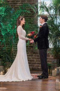 Bride in white dress and groom in black suit smile, holding hands in an indoor venue with plants, candles, and brick walls.