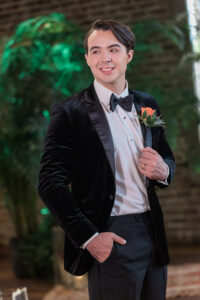 Man in black velvet tuxedo with boutonnière smiles indoors, hand in pocket, with green plants and brick wall behind him.