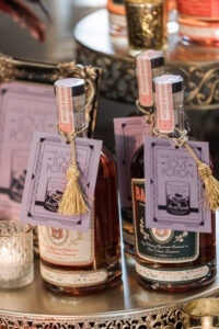 Liquor bottles labeled Hazel & Emmetts Love Potion on an ornate tray with candles and elegant glassware.