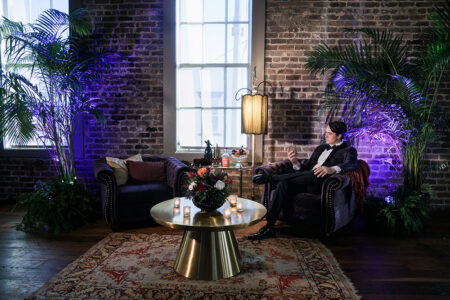 Man in formal wear sits on a dark armchair in a stylish lounge with plants, brick walls, and a gold coffee table with flowers.