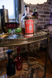 Backyard whiskey bottle on brass bar cart with vintage glassware, decanters, and shaker by brick wall and window.