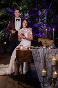 Bride in white and groom in red jacket toast at lace table, smiling amid greenery and purple lighting.