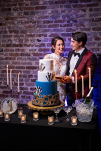 A formally dressed couple stands by a geometric cake, ready to cut it, with candles and plates on a table against a brick wall.