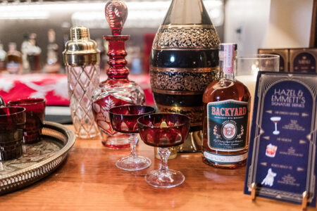 Bar setup with red crystal glasses, Backyard Strawberry Rye Whiskey, ornate decanters, drink menu, and shaker on wood counter.