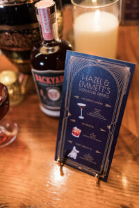 Hazel & Emmetts blue cocktail menu on wood table with liquor bottle, red drink, gold cup, and illustrated drink icons.