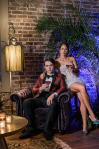 Man in red tuxedo holding a drink sits in velvet chair; woman in silver dress stands beside him, arm on his shoulder.