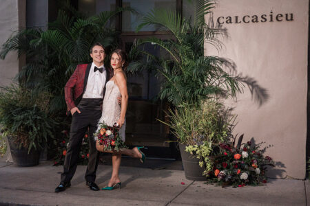 A well-dressed couple stands outside Calcasieu, surrounded by potted plants and a flower arrangement.