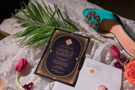 Hazel & Emmett wedding invitation with gold accents on lace, surrounded by flowers, palm leaf, envelope, and jeweled teal shoe.