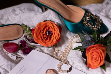 Teal jeweled heels, orange roses, pearls, and a cameo necklace on lace with an open envelope in a romantic flat lay.