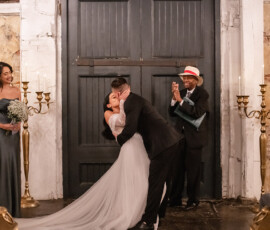 Bride and groom kiss indoors at their wedding as smiling guests and wedding party applaud in a joyful, candlelit setting.