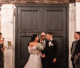 Bride and groom smiling, holding hands indoors at their wedding ceremony with an officiant, guests, lanterns, and candles.
