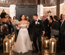 Smiling couple walks hand-in-hand down the aisle as guests clap in a warmly-lit, lantern-decorated wedding venue.