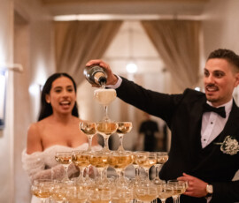Man in tuxedo pours champagne into tower as woman in white dress smiles beside him in an elegant, warmly lit room.