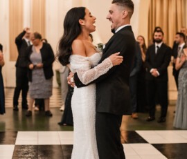 Bride and groom smile during their first dance on a checkered floor as guests celebrate in an elegant ballroom.
