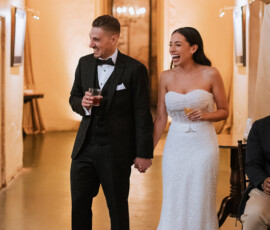 Bride in strapless gown and groom in black tux hold hands, laughing with drinks near indoor draped curtains.