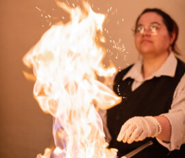 Chef with glasses and gloves flambés food in a pan, sending tall flames into the air in the background.