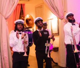 Three musicians in white shirts clap while holding brass instruments indoors; a sousaphone player stands behind them in vibrant lighting.