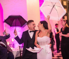 Bride and groom dance with black and white parasols, surrounded by guests at a colorful indoor wedding reception.