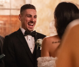 A groom in a tuxedo laughs joyfully at his bride during a warmly lit wedding ceremony.