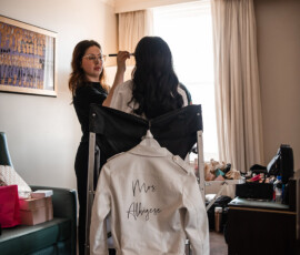 A woman has her makeup done in a bright room; a “Mrs. Albigese” jacket hangs on a chair amid makeup and gifts.