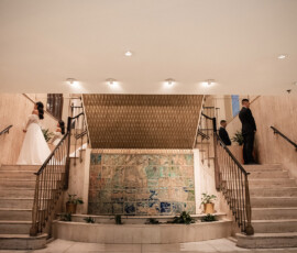Bride and groom ascend separate marble staircases in a modern venue with a central tiled mural and plants.