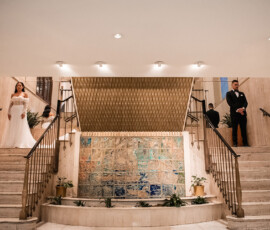 Bride and groom stand separately on marble staircases in an elegant venue with a tiled mural and gold accents.