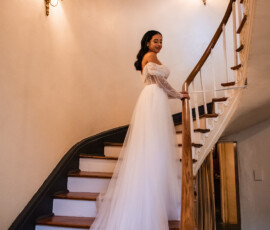 Bride in off-the-shoulder gown with long train stands on curved staircase, softly lit by wall sconces.