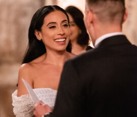 Smiling bride in off-shoulder dress faces groom holding handwritten vows at indoor wedding ceremony.