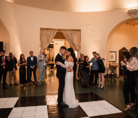 Bride and groom share their first dance in a warmly lit room as guests watch, smile, and record the moment on their phones.