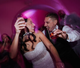 Bride and groom laughing and dancing with drinks at their wedding, pink lights and celebrating guests in the blurred background.
