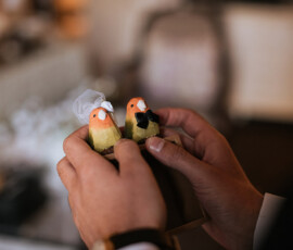 Hands holding bird bride and groom figurines with veil and bow tie, against a softly blurred background.