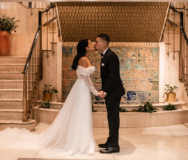 Bride and groom kiss, holding hands indoors near marble stairs and decorative plants.