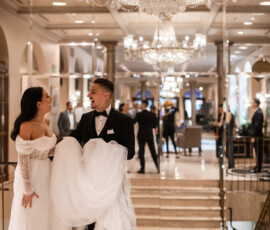Bride and groom smiling under chandeliers in a marble hall, the groom lifting part of the bride’s dress in formal wedding attire.