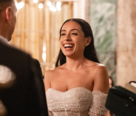 Bride in an off-the-shoulder white dress holds hands with partner at a warmly lit wedding ceremony.