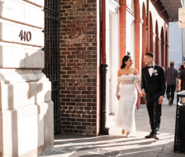 Bride and groom in sunlight walk hand-in-hand on a city sidewalk, smiling; street number 410 is visible.