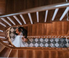 Bride and groom embracing at the base of a wooden spiral staircase, seen from above on a checkered tile floor.