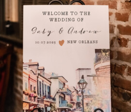 Wedding welcome sign on easel reads: Welcome to the wedding of Gaby & Andrew, 10.07.2023, New Orleans, with colorful buildings illustration.