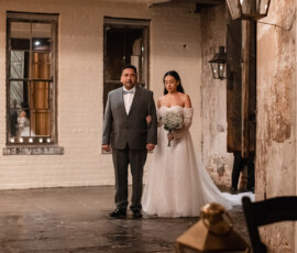Bride in white gown with flowers stands arm-in-arm with man in suit indoors, ready to walk down the wedding aisle.