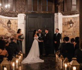 Bride and groom hold hands at indoor ceremony, with guests, candles along the aisle, officiant, and attendants nearby.