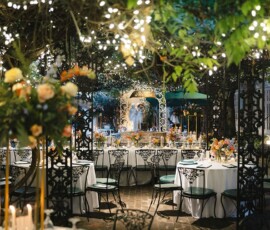Outdoor wedding with round tables, floral centerpieces, iron chairs, string lights, and a bride and groom under a lush arch.