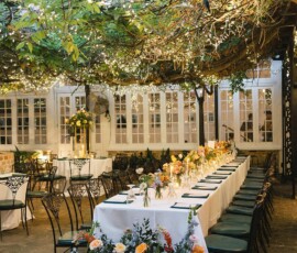 Outdoor dining area with flower-adorned long tables, black chairs, string lights, greenery, and a large floral arrangement.