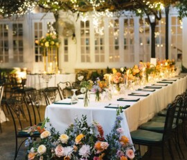 Garden wedding table with white cloth, floral centerpieces, and candles under string lights; chairs await guests.
