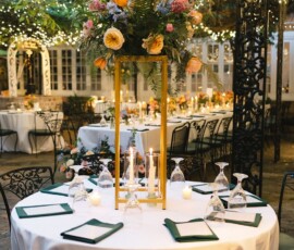 Outdoor round table with green chairs, floral centerpiece, candles, menus, and wine glasses under romantic string lights.