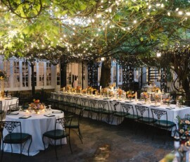 Outdoor wedding reception with long tables, floral centerpieces, string lights, and wrought iron chairs under leafy garden trees.