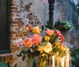 Floral arrangement with roses and greenery on a wooden table, candles lit, brick wall and plants in the background.