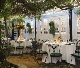Charming outdoor patio with round tables, white linens, black chairs, floral centerpieces, string lights, and greenery overhead.