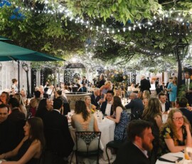 Formally dressed guests sit at round tables under string lights at an elegant, greenery-filled Gulf South-inspired outdoor event.