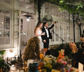 Bride and groom at outdoor New Orleans wedding; groom toasts as guests clap under string lights and floral decorations.