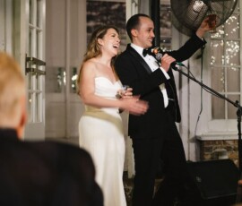 Bride and groom in formalwear smile and hold drinks as the groom speaks into a mic at an indoor New Orleans wedding.